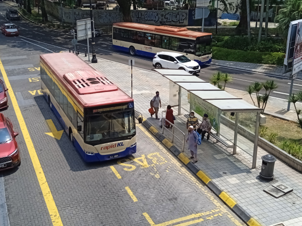 Stadtstraße mit fahrenden Autos, Bussen, Bushaltestelle, Fußgängern auf dem Gehweg, Strommasten, Beschilderung, Bäumen, Grünflächen und umliegenden Gebäuden.