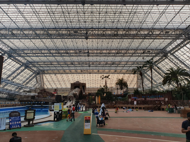 Eine Gruppe von Menschen, die um ein Hallenbad mit Bäumen, Informationsschildern und anderen Objekten in einem Sommerland-Freizeitpark herumstehen und sitzen, unter einer Decke mit Stangen und einem klaren blauen Himmel im Hintergrund.