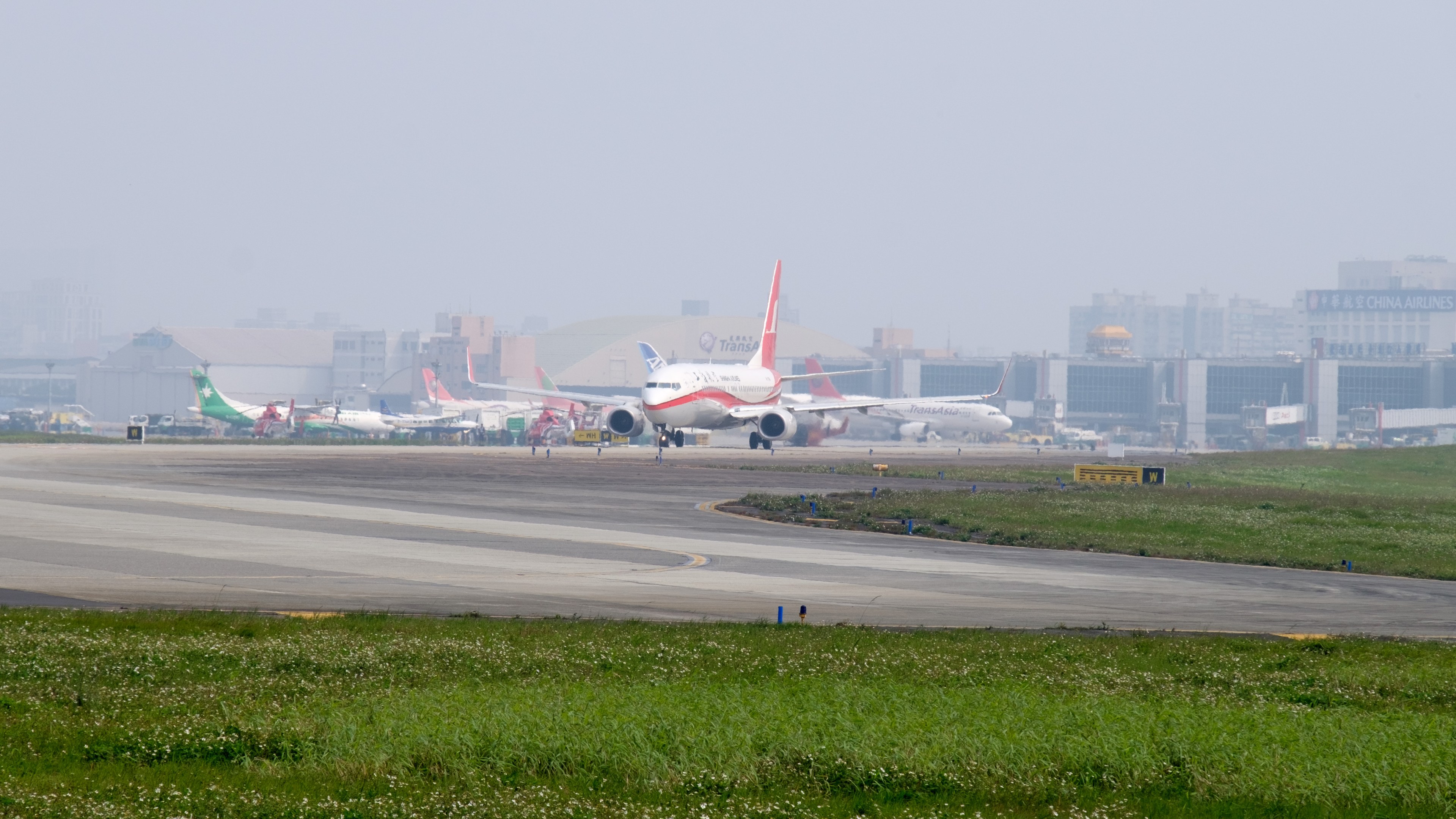 Flughafenvorfeld mit mehreren geparkten Flugzeugen, grünem Umfeld, Gebäuden im Hintergrund und einem klaren blauen Himmel.