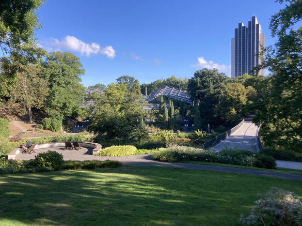 Ein Park mit grünem Gras, Bäumen, Pflanzen, Blumen, Bänken und einem Weg, mit Gebäuden und einem bewölkten Himmel im Hintergrund.