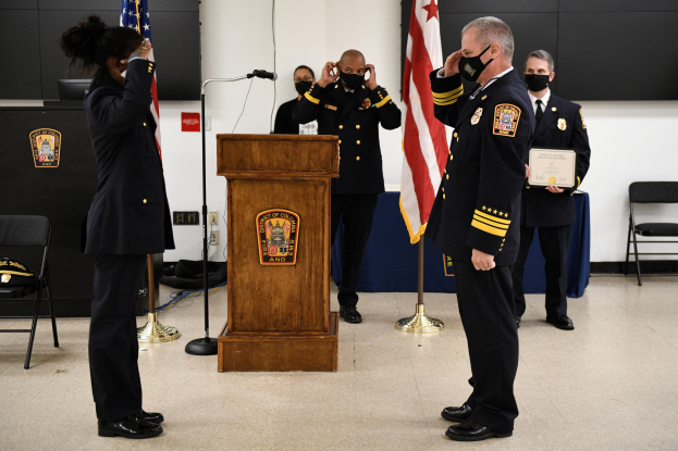 Gruppe uniformierter Personen in Masken vor einem Rednerpult bei einer Vereidigung eines neuen Feuerchefs, der eine Urkunde hält, mit Flaggen, Stühlen, Tischen und einer Wand im Hintergrund.