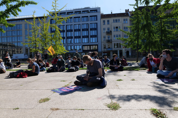 Menschen sitzen vor einem Gebäude auf dem Boden während einer Demonstration in Berlin, einige tragen Masken und sind von Taschen und Bäumen umgeben unter einem klaren blauen Himmel.