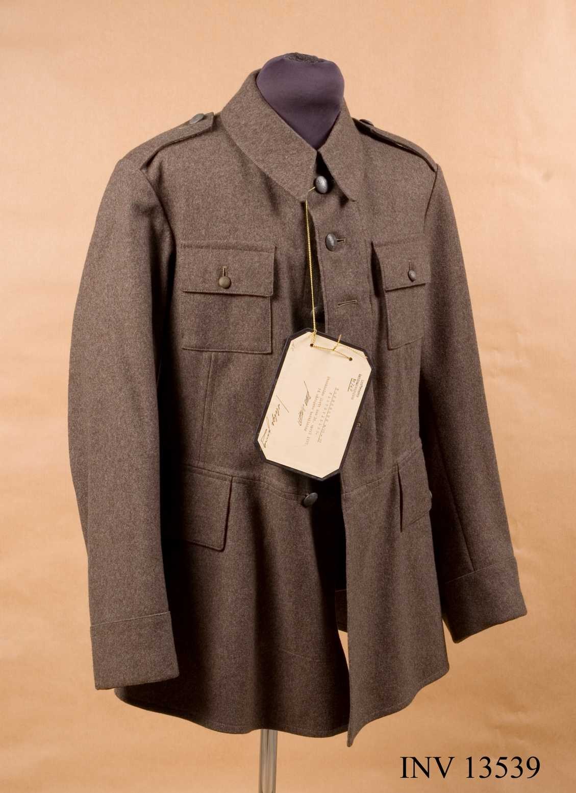 Luftwaffen-Offiziersjacke der deutschen Wehrmacht mit einem Etikett, das an einer Schaufensterpuppe gegen einen cremefarbenen Hintergrund gehängt ist, begleitet von Text unten.