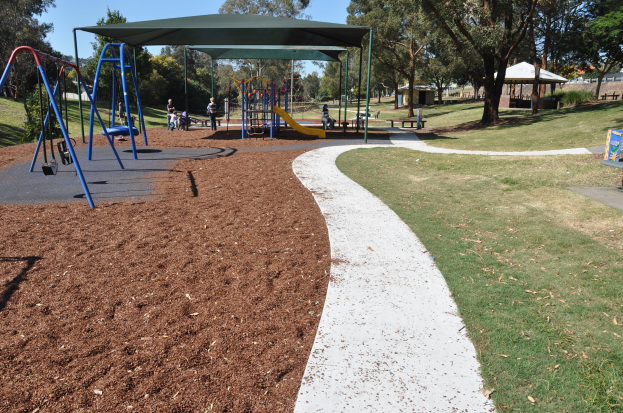 Ein Park mit einem Spielplatz mit Schaukeln, Rutschen und anderen Spielgeräten, umgeben von grünem Gras, Bäumen, Bänken, Schuppen im Hintergrund und einem klaren blauen Himmel.