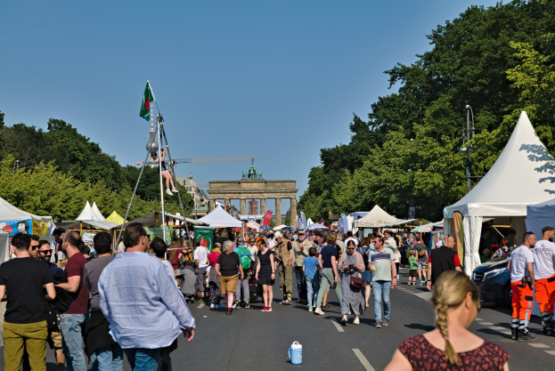 Eine Menschenmenge geht eine Straße entlang, gesäumt von Zelten, Fahrzeugen und Bäumen, auf einen Bogen unter einem klaren blauen Himmel zu, mit Fahnenmasten auf der linken Seite, was auf das Oktoberfest in München, Deutschland, hindeutet.