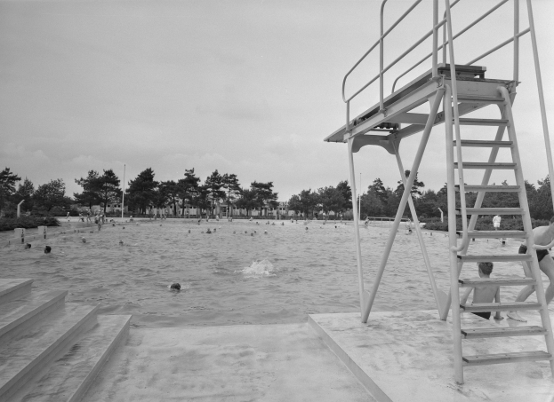 Schwarzes und weißes Foto von Menschen, die im Wasser eines Strandes schwimmen, mit einem Rettungsturm auf der rechten Seite, Bäumen und Pfählen im Hintergrund und einem klaren Himmel.