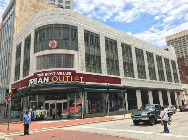 Eine belebte Stadtstraße mit Fahrzeugen und Fußgängern, hohen Glasgebäuden und einem Schild mit der Aufschrift "Der beste Preis für städtische Ausgänge" unter einem bewölkten Himmel.