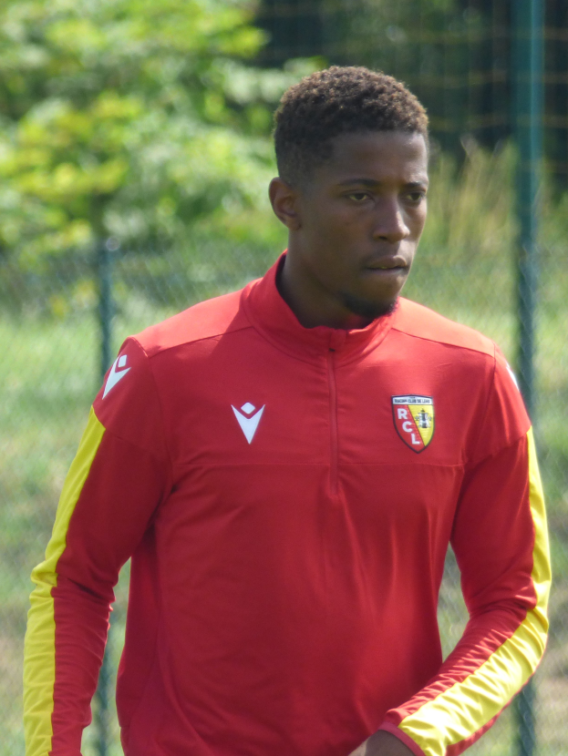 Ein junger Mann in rot-gelbem Trikot steht auf einem Fußballfeld, umgeben von einem Zaun und Bäumen, mit entschlossener Miene, wahrscheinlich ein Flügelstürmer.