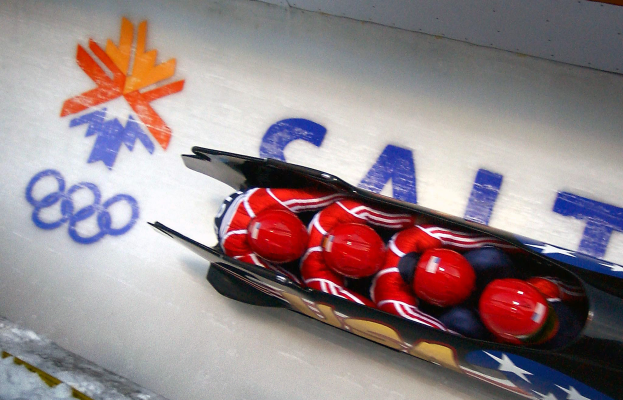 Ein Bobfahrer bei den Olympischen Spielen, mit einem Fahrzeug im Vordergrund und einer Wand mit Text und einem Gemälde im Hintergrund.