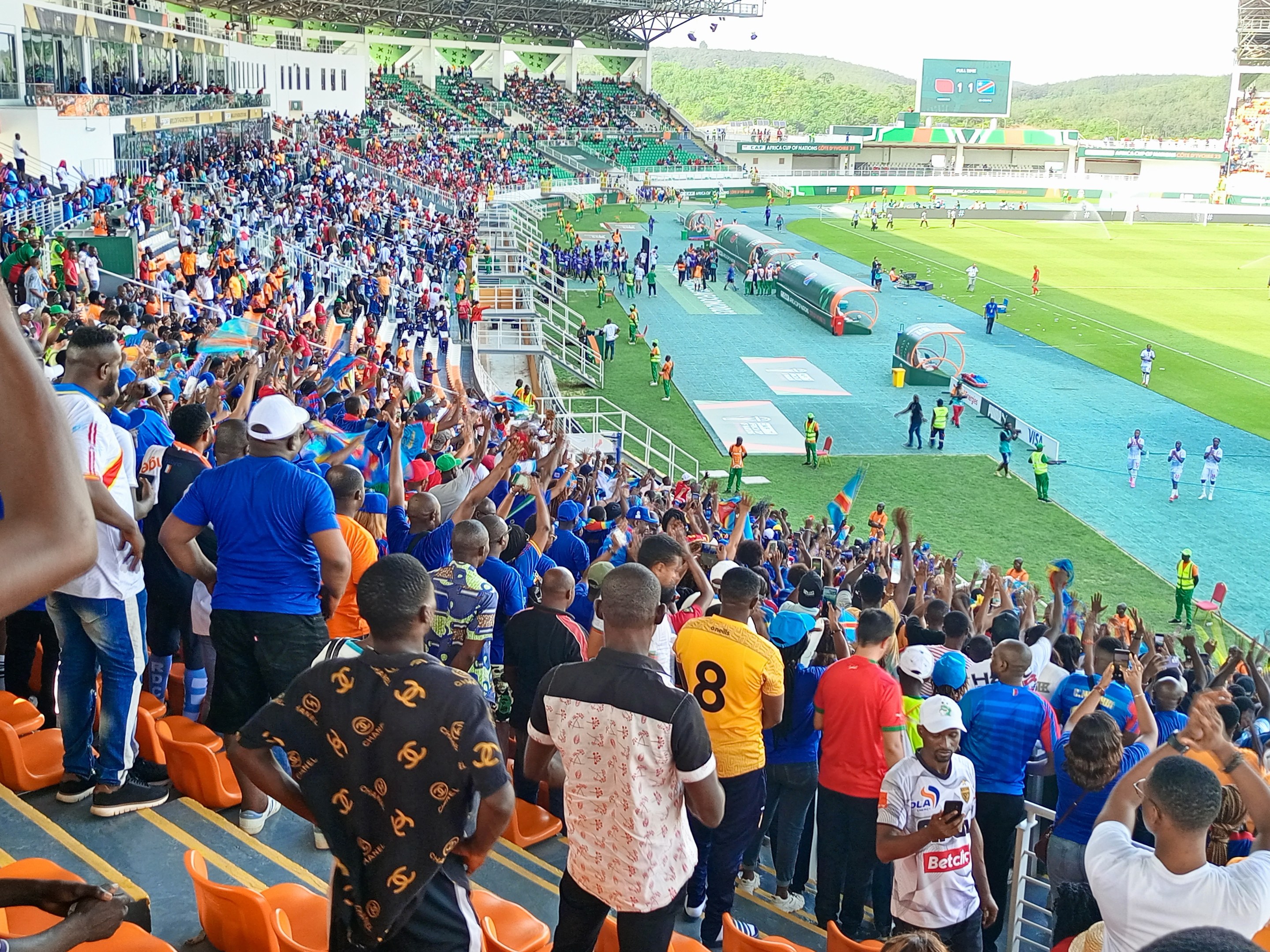 Eine große Menschenmenge versammelt sich vor einem eingezäunten Fußballfeld mit Hügeln und einem klaren blauen Himmel im Hintergrund und wartet auf den Beginn eines Spiels während der 2019 African Cup of Nations.
