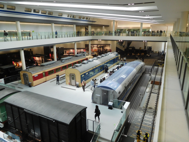 Eine große Museumsausstellung zeigt verschiedene Züge in unterschiedlichen Größen und Farben, mit Menschen, die von verschiedenen Ebenen und Geländern aus beobachten, unter Deckenbeleuchtung.