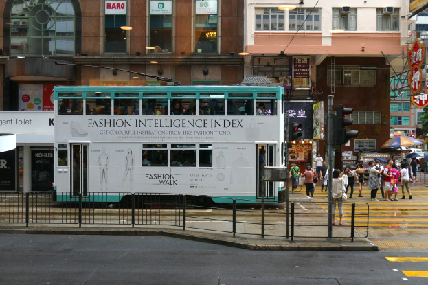 Ein Doppeldeckerbus fährt eine Stadtstraße entlang mit hohen Gebäuden, Fußgängern auf dem Bürgersteig mit Regenschirmen, Verkehrsampeln, einer Straßenreling, Informationsschildern, Bäumen und einem klaren blauen Himmel.