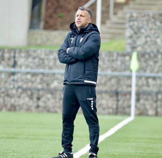 Ein Mann in schwarzer Jacke und Hose steht auf einem Fußballfeld mit verschränkten Armen, der wie ein Trainer während eines Trainings aussieht, mit einem Zaun, einer Treppe und einer Wand im Hintergrund.