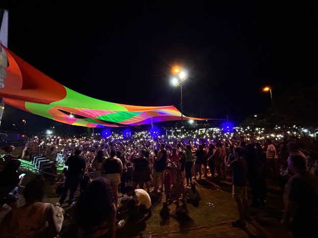Eine große Menschenmenge steht vor einem hell erleuchteten Zelt in der Nacht, umgeben von farbigen Lichtern und beleuchteten Pfählen, einem Gebäude auf der linken Seite und dem Nachthimmel im Hintergrund.