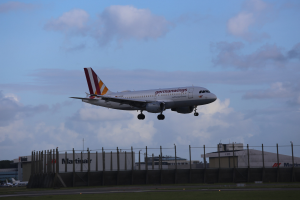 Germanwings Airbus A320-200 beim Start von Frankfurt Airport mit Gras, einem Zaun, Gebäuden, Bäumen und einem bewölkten Himmel im Hintergrund.