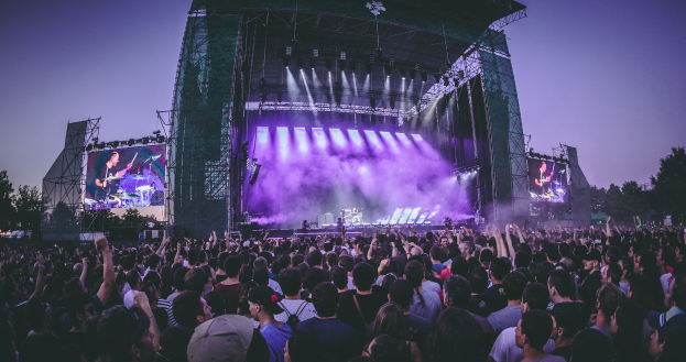 Eine große Menschenmenge versammelt sich auf einem Musikfestival mit einer Bühne, auf der Musiker spielen, die von Scheinwerfern beleuchtet und von Bildschirmen umgeben sind, vor einer Kulisse aus einem Gebäude, Bäumen und einem klaren blauen Himmel.