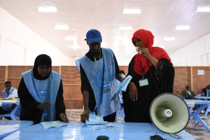 Eine Gruppe von Menschen steht um einen Tisch mit einem Megafon, Papieren und anderen Gegenständen herum, während andere im Hintergrund auf Stühlen oder stehen, unter einer Decke mit Lampen, was an eine Wahllokal erinnert.