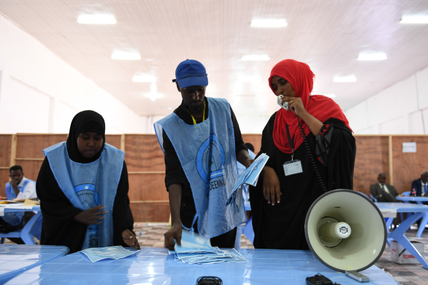 Eine Gruppe von Menschen steht um einen Tisch mit einem Megafon, Papieren und anderen Gegenständen herum, während andere im Hintergrund auf Stühlen oder stehen, unter einer Decke mit Lampen, was an eine Wahllokal erinnert.