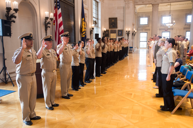 Eine Gruppe uniformierter Personen steht in einem Raum mit Stühlen auf der rechten Seite, Fotorahmen an der Wand, Kerzen, einem Lautsprecher auf der linken Seite und einer Fahne in der Mitte, alle salutierend während einer Militär-Offizierszeremonie.