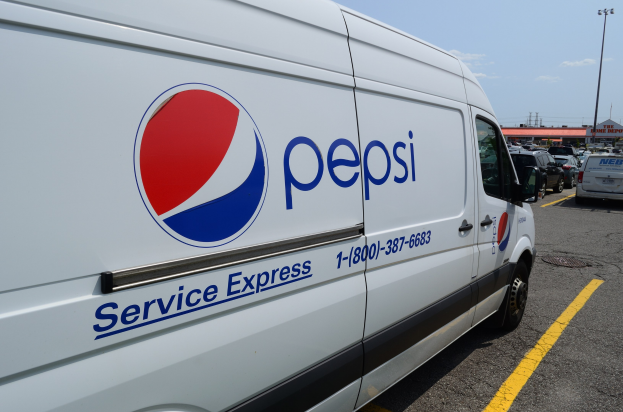 Pepsi Service Express Lieferwagen auf einem Parkplatz mit anderen Fahrzeugen, Laternen, Gebäuden, Bäumen und einem klaren blauen Himmel im Hintergrund.