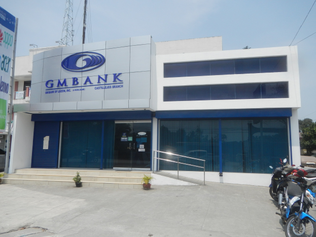 Außenansicht der Gmbank Bank in Hyderabad mit Glaswänden, Treppen mit Geländern, Topfpflanzen, einer Straße mit Fahrzeugen, einem Banner, einem Turm mit Drähten, Bäumen und einem bewölkten Himmel.