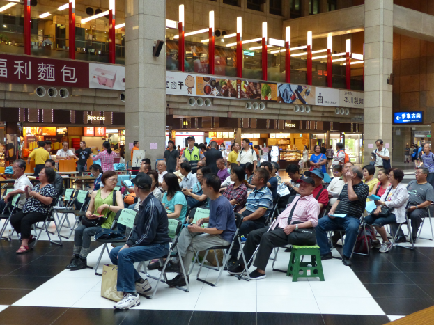 Eine große Gruppe von Menschen sitzt und steht in einem Flughafen, mit Gägen auf Hockern, Schildern, Säulen, einem Anzeigebildschirm, Deckenleuchten und einem Gebäude im Hintergrund, wahrscheinlich an einer Messeveranstaltung teilnehmend.