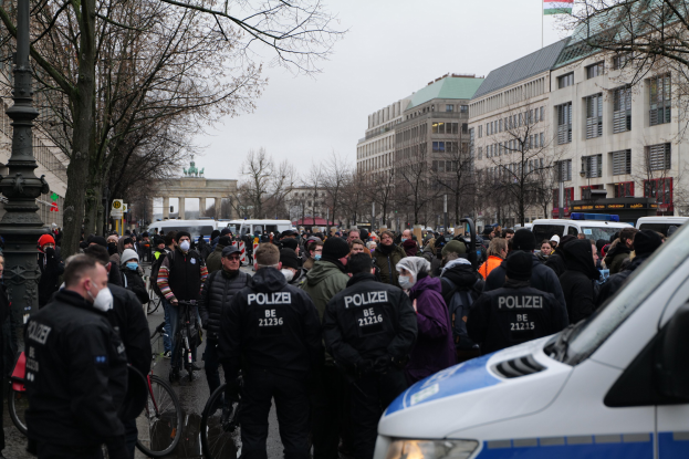Große Gruppe von Menschen vor einem Polizeibus auf einer Straße mit Bäumen, Laternen und Gebäuden, einige tragen Mützen und Masken, Fahrräder im Vordergrund, Bogen mit Säulen und Flagge im Hintergrund unter sichtbarem Himmel.
