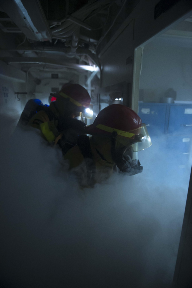 Zwei Feuerwehrleute in Helmen und Schutzausrüstung stehen in einem dunklen, rauchigen Raum mit einer Tür auf der rechten Seite und Rohren im Hintergrund.