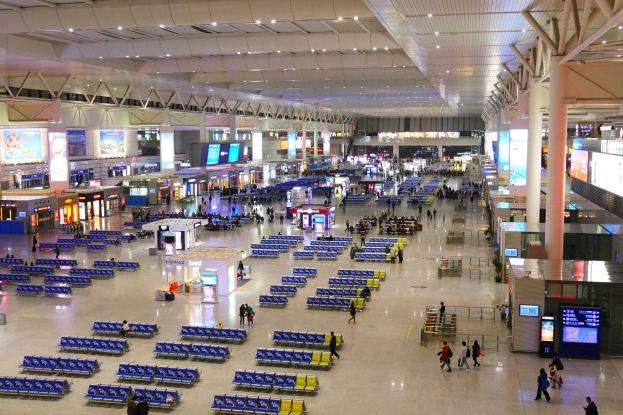 Innensicht eines Flughafen-Terminals mit Menschen, blauen Stühlen, Geschäften, Informationsanzeigen, Bildschirmen, tragenden Säulen, Deckenbeleuchtung und verschiedenen Einrichtungsgegenständen.