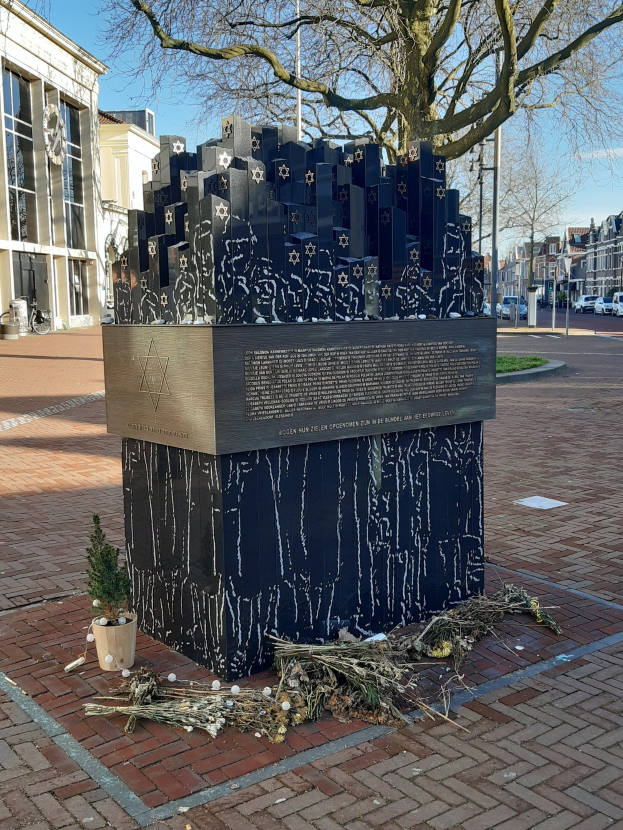 Denkmal in einem Stadtplatz für Holocaust-Opfer, mit einem Baum davor, einem Topfgewächsz auf dem Boden, verstreuten trockenen Blättern und Gebäuden, Fahrzeugen, Masten und Bäumen im Hintergrund.
