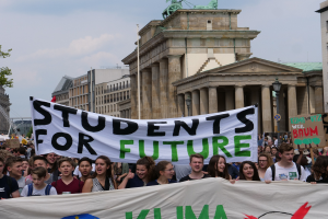 Eine Gruppe von Schülern marschiert in Berlin, die eine bunt bemalte "Students for Future"-Fahne trägt, vor einem Hintergrund aus Gebäuden, Bäumen und Himmel.