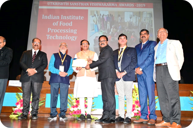 Eine Gruppe von Männern auf einer Bühne mit einem Zertifikat und einem Bildschirm im Hintergrund mit der Aufschrift "Indian Institute of Food Processing Technology Awards 2019" und Blumenschmuck.