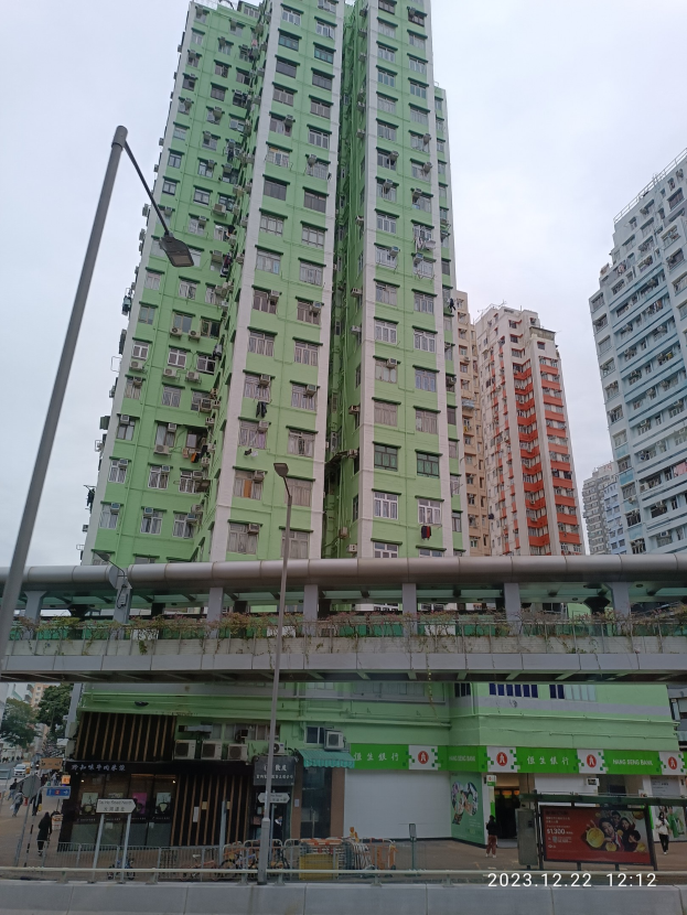 Ein hohes grünes Gebäude mit zahlreichen Fenstern, das sich in einer urbanen Gegend befindet und von anderen Gebäuden, Straßeninfrastruktur, Fahrzeugen, Bäumen und einem bewölkten Himmel umgeben ist.