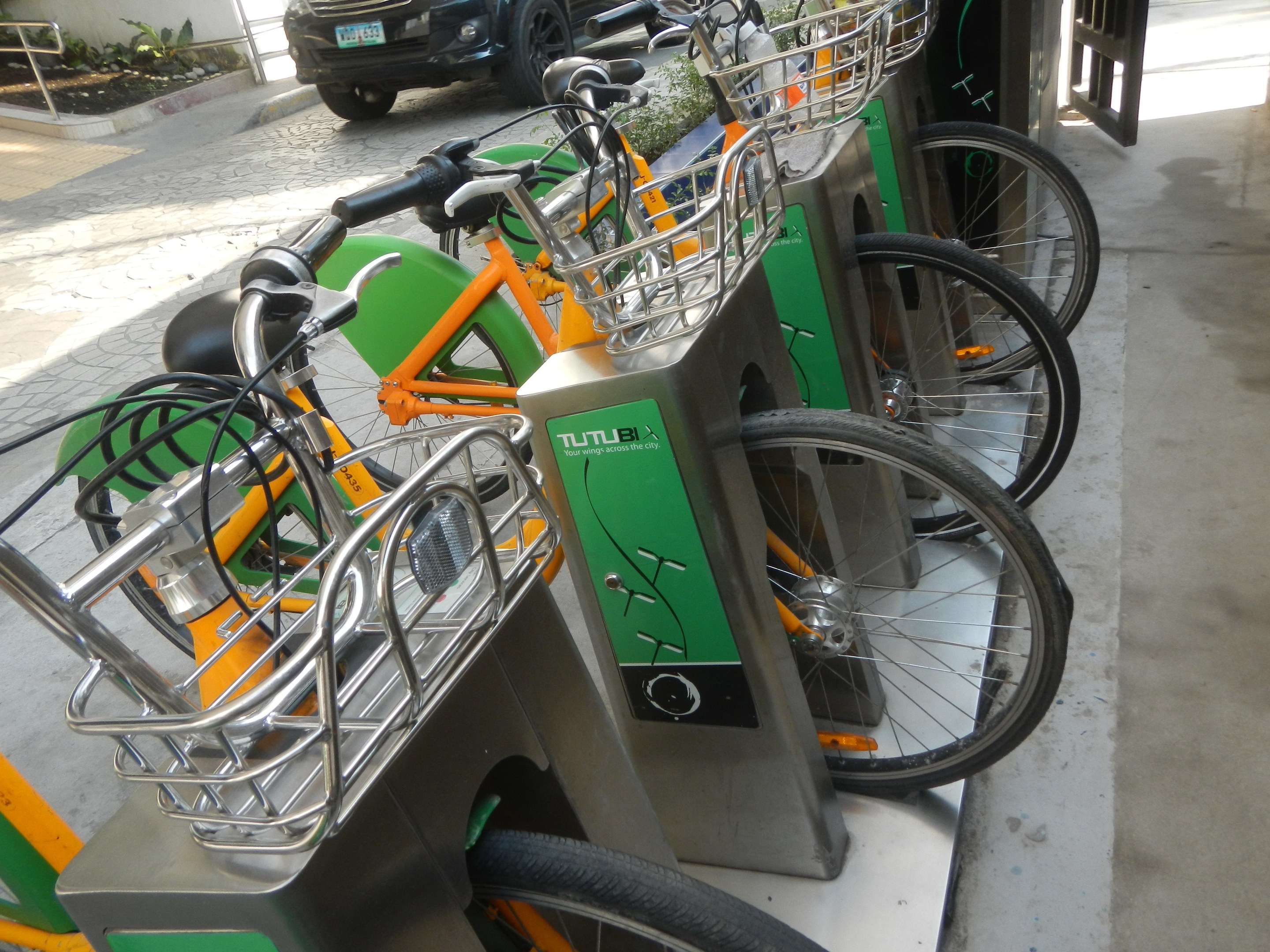 Eine Reihe von Fahrrädern aus einem Shuttle-Fahrradverleihsystem, die in einem Parkplatz geparkt sind, mit einem Auto auf der Straße dahinter, einem Tor auf der rechten Seite und Pflanzen und einer Wand auf der linken Seite.