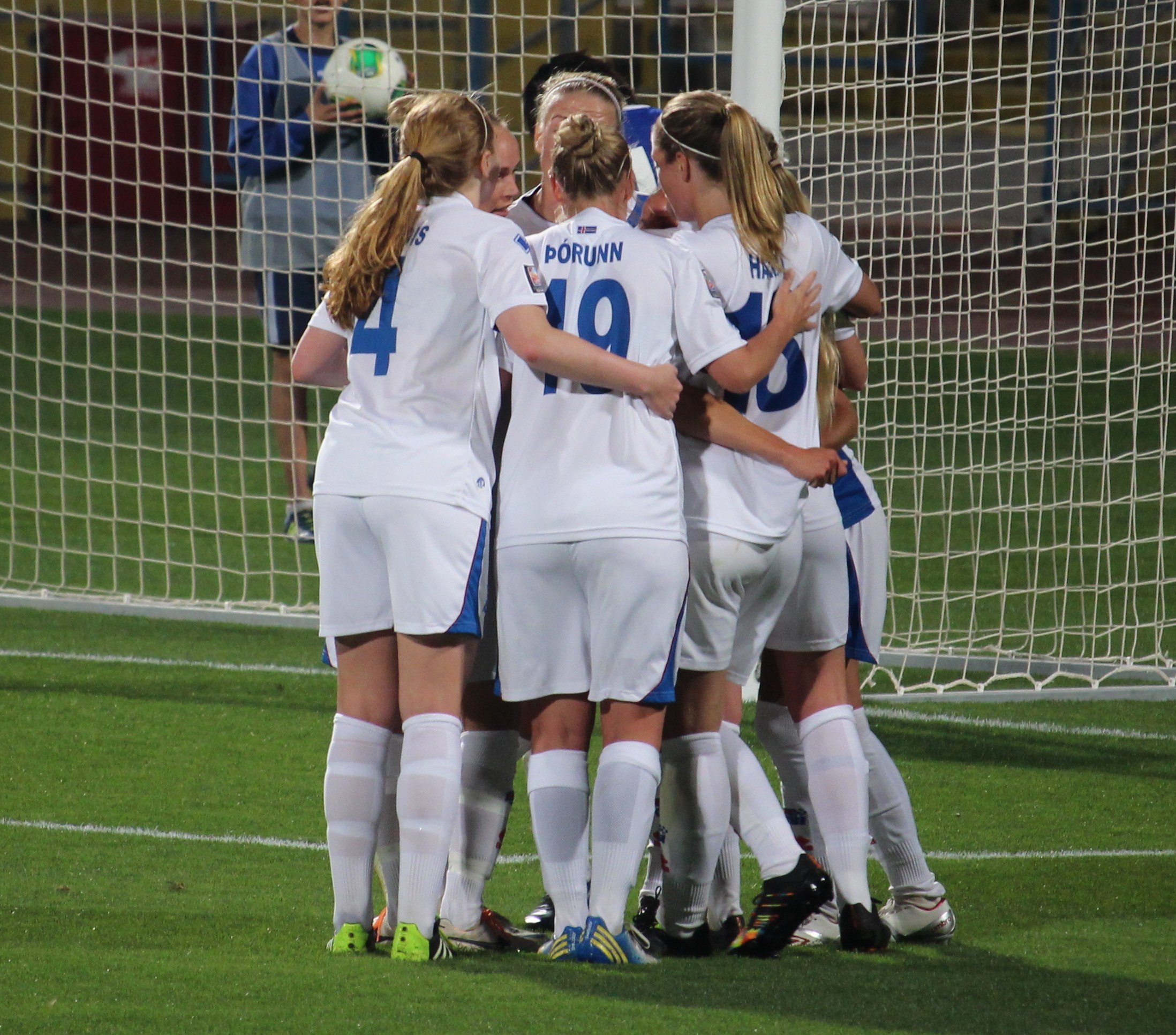 Eine Gruppe von Fussballspielerinnen in Uniformen auf dem Feld mit einem Netz und einer Person, die einen Ball hält, im Hintergrund.