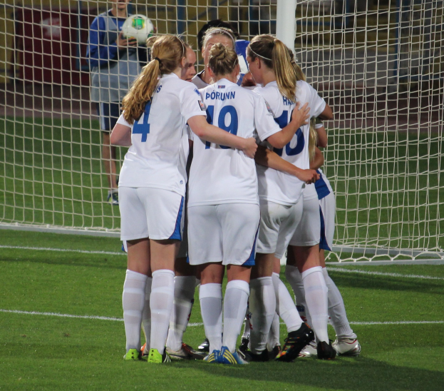 Eine Gruppe von Fussballspielerinnen in Uniformen auf dem Feld mit einem Netz und einer Person, die einen Ball hält, im Hintergrund.