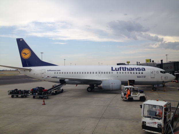 Ein Lufthansa Airbus A320-200 steht auf dem Frankfurter Flughafen, umgeben von verschiedenen Bodenfahrzeugen, Wagen, Verkehrskegeln, Pfosten und Texttafeln unter einem bewölkten Himmel.