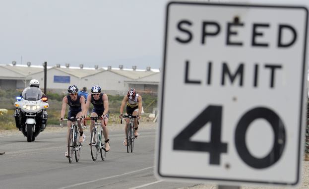 Eine Gruppe von Fahrradfahrern mit Helmen fährt auf einer Straße neben einem Geschwindigkeitsbegrenzungsschild, mit Gebäuden, Versorgungsmästen, Vegetation und einem klaren Himmel im Hintergrund.