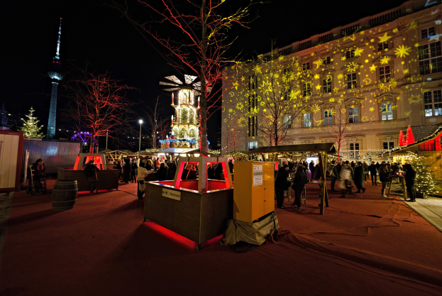 Ein geschäftiger Weihnachtsmarkt in Berlin, Deutschland mit Menschen um dekorierte Stände, festliche Lichter, Bäume, Gebäude, Laternenmasten und einen Turm unter einem dunklen Himmel.