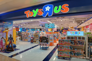 Ein Toys R Us Geschäft in einem Einkaufszentrum, das Regale mit Spielzeug und anderen Artikeln zeigt, ein sichtbares Ladenschild, Deckenbeleuchtung und ein Wasserzeichen in der rechten unteren Ecke.