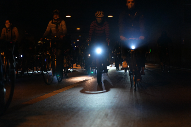 Eine Gruppe von Menschen, die nachts mit Fahrrädern auf einer gut beleuchteten Stadtstraße fahren und alle Helme tragen.