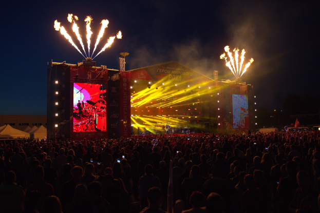 Menschenmenge auf einem Musikfestival mit einer Bühne, auf der Musiker unter hellen Scheinwerfern spielen, links Banner und Zelte, sowie Feuerwerk am dunklen Himmel.