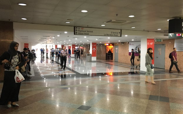 Menschen mit Taschen durch eine Flughafenhalle gehen, mit Hinweistafeln, Deckenlampen und einem Mülleimer im Hintergrund; eine Frau trägt Schuhe.