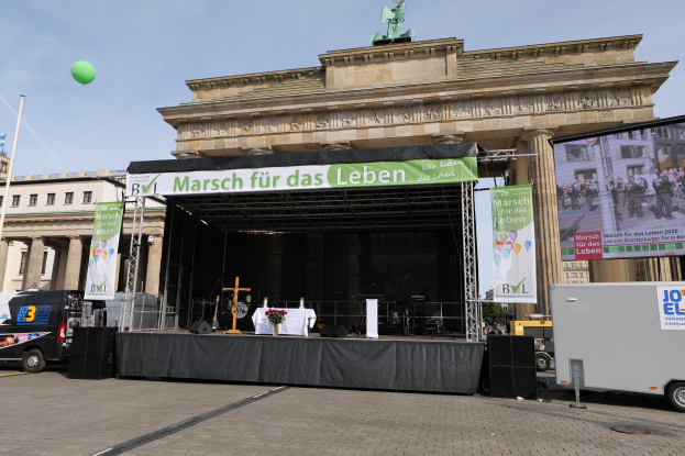 Bühne vor dem Brandenburger Tor mit einem Tisch, Bannern, Lautsprechern, Fahrzeugen, Gebäuden, einer Statue, einer Fahne und Wolken im Hintergrund.