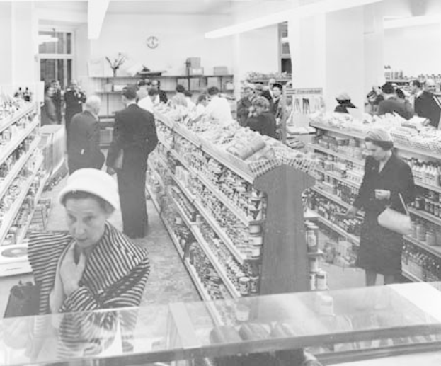 Schwarzes und weißes Foto von Menschen beim Einkaufen in einem Supermarkt mit Regalen voller Lebensmittel, einige halten Taschen, eine Uhr an der Wand und Deckenbeleuchtung.