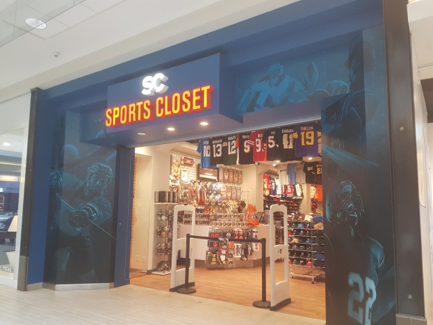 Ein Sportbeköstedt in einem Einkaufszentrum mit einem "SC Sports Closet"-Schild, das Sportbeköbung an Bügeln und organisierte Gegenstände in Schränken unter Deckenlampen zeigt, mit einer gläsernen Eingangstür auf der linken Seite.