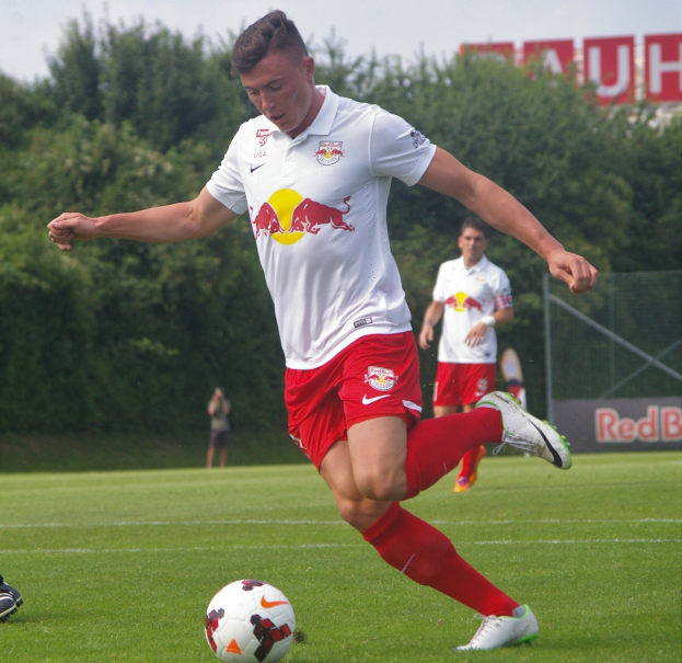 Ein professioneller Füßballspieler kickt einen Ball auf einem grünen Feld mit Bäumen und einem klaren blauen Himmel im Hintergrund, in der Nähe einer Tafel mit der Aufschrift "RB Leipzig."