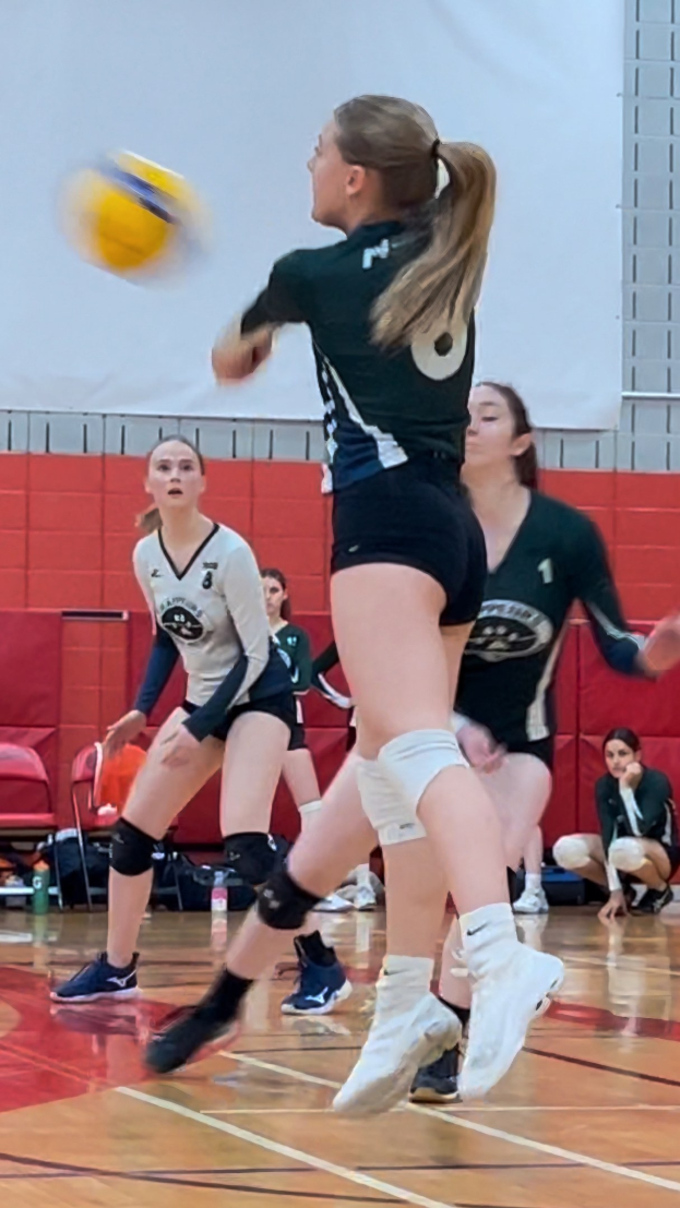 Eine Gruppe junger Frauen beim Volleyballspielen mit einem Ball in der Luft, eine Wand, Stühle, Taschen, Flaschen und Turnschuhe im Hintergrund.
