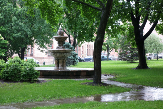 Ein Brunnen in einem grünen Park mit Bäumen, Pflanzen und Gras, umgeben von Fahrzeugen auf einer Straße, mit Gebäuden und einem blauen Himmel im Hintergrund.