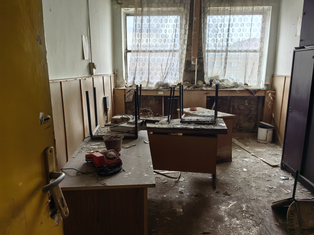 Leerer Raum mit einer Tür auf der linken Seite, einem Schrank auf der rechten Seite und einem Fenster mit Vorhängen im Hintergrund; ein zentraler Tisch hält verstreute Büromaterialien.